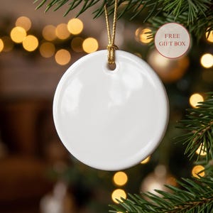 May include: A round, white ceramic ornament with a smooth, glossy finish. The ornament features a small hole at the top, through which a thin, gold-colored string is looped for hanging. The surface of the ceramic disc is entirely blank.