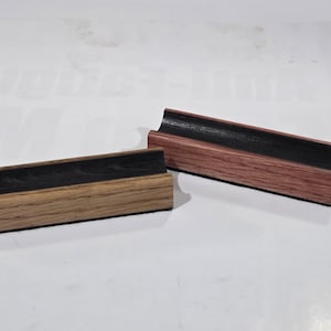 May include: Two rectangular wooden pen rests with a concave top surface. One rest features a light brown wood grain base with a black interior, while the other has a reddish-brown wood grain base with a black interior, both showing wood texture.