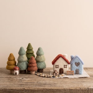 Op de afbeelding: Een verzameling van naaldgevilte items, waaronder vijf groenblijvende bomen in tinten groen, geel en roest, een kleine paddenstoel, twee miniatuurhuisjes met een rood en blauw dak, een piepklein houten bankje en een pad van kleine grijze steentjes.