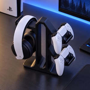 May include: A black gaming accessory stand featuring a central vertical support with arms to hold one pair of white and black over-ear headphones on the left side and two white and black wireless game controllers on the right side.