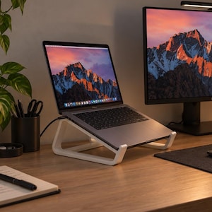 May include: A white laptop stand holding a silver laptop on a wooden desk. The setup includes a large computer monitor with a mountain landscape wallpaper, a black keyboard, a computer mouse, a notebook with a pen, and a black water bottle.