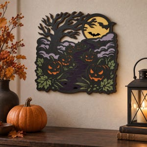 Op de afbeelding: Een gelaagd houten wandkunstwerk met een Halloween-tafereel, bestaande uit een zwarte boom, een gele maan, vliegende vleermuizen en diverse uitgesneden pompoenen met oranje ogen en monden tegen een achtergrond van paars en groen gebladerte.