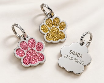 May include: Three paw-shaped pet ID tags with silver-toned metal edges and split rings. One tag features a pink glitter inlay, one features a gold glitter inlay, and one is plain silver with the engraved text SIMBA and 07700 900123.