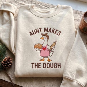 May include: A cream-coloured crewneck sweatshirt featuring an embroidered graphic of a goose wearing a pink apron and a brown headscarf. The goose holds a loaf of bread and a rolling pin. The text reads AUNT MAKES THE DOUGH.