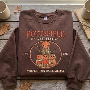 Puede incluir: Una sudadera marrón de cuello redondo con un bordado del Pottsfield Harvest Festival. El diseño incluye una figura central con cabeza de calabaza rodeada por tres personajes de calabaza más pequeños, con el texto "POTTSFIELD HARVEST FESTIVAL", "EST. 1502" y "YOU'LL JOIN US SOMEDAY".