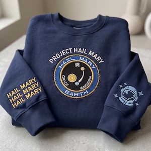 May include: A navy blue crewneck sweatshirt featuring an embroidered circular patch on the chest with the text Project Hail Mary, Earth, and Grace. The left sleeve has the text Hail Mary repeated in yellow, and the right sleeve has an astronaut helmet.