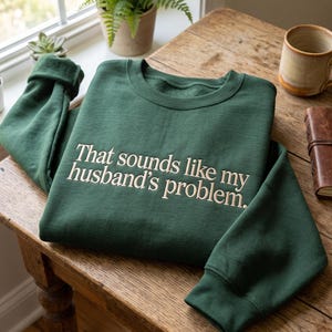 Puede incluir: Una sudadera de cuello redondo en color verde bosque oscuro con el texto bordado "That sounds like my husband's problem." en una fuente serif de color beige claro. La prenda tiene mangas largas con puños y cuello acanalados.