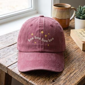 May include: A dusty rose baseball cap featuring embroidered white text that reads "hoa hoa hoa hoa" arranged in a slight arc, accented by several small, four-pointed yellow stars stitched above and around the lettering on the front panel.