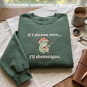 May include: A forest green crewneck sweatshirt featuring an embroidered graphic of a light green frog with a brown mushroom cap playing a small guitar. The text reads, "If I shenan once... I'll shenanigan." The sweatshirt has long sleeves and ribbed cuffs.