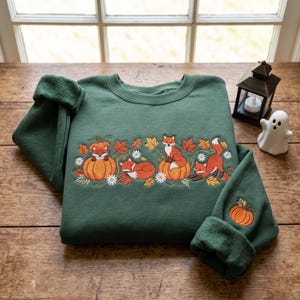 Könnte beinhalten: Ein dunkelgrüner Rundhals-Pullover mit einer Stickerei von drei orangefarbenen Füchsen, Kürbissen, Herbstblättern und weißen Gänseblümchen auf der Brust. Ein kleiner orangefarbener Kürbis ist zudem auf dem unteren Teil des rechten Ärmels gestickt.