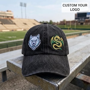 May include: A charcoal gray baseball cap featuring an embroidered white and light blue wolf head on the left panel and a green and gold dragon emblem on the right panel. The text CUSTOM YOUR LOGO is visible in the upper right corner of the image.