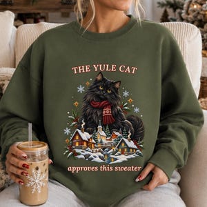 May include: An olive green crewneck sweatshirt featuring a graphic of a fluffy black cat wearing a red patterned scarf. Below the cat is a snowy village scene with houses and trees. The text reads THE YULE CAT approves this sweater.