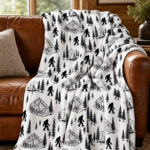 May include: A white fleece throw blanket featuring a repeating black graphic print of Bigfoot silhouettes, pine trees, mountain ranges, and small camping tents. The soft fabric displays a detailed wilderness pattern across the entire surface.