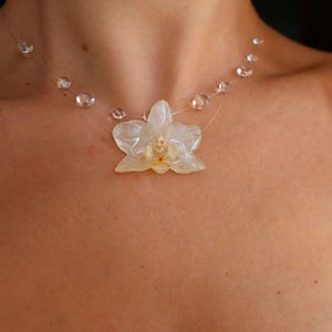 Puede incluir: Un collar con un colgante translúcido en forma de orquídea de color amarillo pálido suspendido de un cordón fino y transparente. El cordón está adornado con pequeñas cuentas redondas y claras espaciadas uniformemente alrededor del cuello.