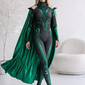May include: A black and green full-body jumpsuit featuring a high collar, cold-shoulder cutouts, and a textured pattern with metallic green accents. The garment includes a long, flowing, metallic green cape attached at the shoulders.