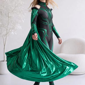 May include: A black bodysuit featuring a high neck, cold-shoulder cutouts, and long sleeves with metallic green geometric patterns. A long, flowing, metallic green cape is attached to the shoulders, creating a dramatic silhouette with a vibrant sheen.
