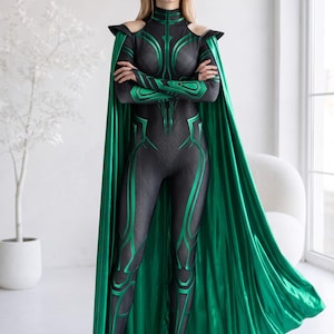 May include: A black and green full-body jumpsuit featuring a high neck, cold-shoulder cutouts, and long sleeves. The garment has a textured pattern with metallic green accents and is paired with a long, flowing, solid metallic green cape.