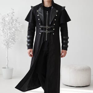 May include: A long black coat featuring a high collar, silver-tone buttons, and a central zipper closure. The garment includes two horizontal buckled straps across the chest, shoulder capes, and three buckled straps on each sleeve cuff.
