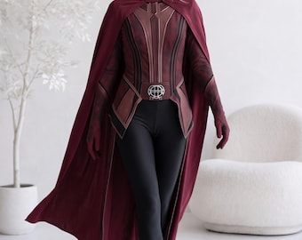 May include: A maroon long-sleeved top with a structured bodice and a matching cape, paired with black leggings. The outfit includes a maroon headpiece with a pointed design and a belt featuring a circular silver buckle with a grid pattern.