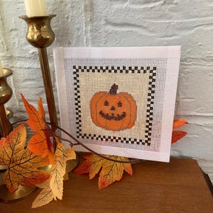 May include: A square needlepoint canvas featuring a stitched orange jack-o'-lantern with a brown stem and a dark brown stitched face. The pumpkin is centred on a cream-coloured background, framed by a black and white checkered border pattern.