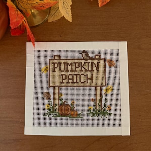 Könnte beinhalten: Ein quadratisches Stickbild auf Stramin mit einem braunen Holzschild, das den Text PUMPKIN PATCH in dunkelbraunen Buchstaben zeigt. Das Motiv enthält einen kleinen Vogel auf dem Schild, einen Kürbis am Fuß sowie grünes Laub mit gelben Blumen.