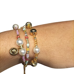 Puede incluir: Tres pulseras apiladas que presentan una mezcla de perlas blancas, pequeñas cuentas de vidrio translúcido multicolor y cuentas metálicas de tono dorado. Una pulsera incluye un pequeño dije redondo y texturizado en forma de girasol.