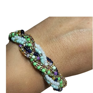 Puede incluir: Una pulsera trenzada construida con múltiples hilos de pequeñas cuentas de vidrio en una variedad de colores que incluyen verde lima, azul claro, blanco, azul marino oscuro, morado y oro metálico, dispuestos en un patrón tejido.