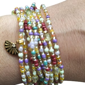 Puede incluir: Una pulsera de múltiples hilos compuesta por numerosas cuentas de vidrio pequeñas y coloridas en tonos amarillo, morado, rojo, turquesa y transparente. La pulsera presenta un pequeño dije de metal dorado en forma de abanico.