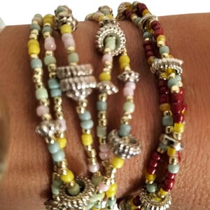 Puede incluir: Una pulsera de múltiples hilos con pequeñas cuentas de vidrio en tonos amarillo, azul claro, rosa y rojo oscuro, intercaladas con cuentas espaciadoras de color plateado y acentos metálicos circulares texturizados en capas.