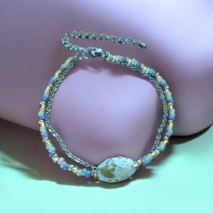 Puede incluir: Una pulsera de doble hilo con una cadena de metal plateado y un hilo de pequeñas cuentas de vidrio multicolor en tonos morado, turquesa, rosa y dorado. Una cuenta ovalada blanca más grande con motas doradas sirve como punto focal.