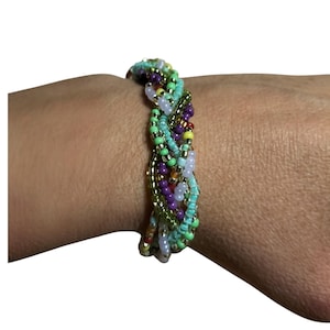 Puede incluir: Una pulsera trenzada de múltiples hilos construida con pequeñas cuentas de vidrio en una paleta vibrante de verde lima, turquesa, morado, amarillo y tonos iridiscentes claros, tejidas para crear una banda circular texturizada y multicolor.
