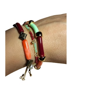 Puede incluir: Un conjunto de tres pulseras superpuestas con una mezcla de cuentas cilíndricas en granate, naranja brillante y verde menta, acentuadas con espaciadores de metal dorado, pequeñas cuentas rojas y verde claro, y una perla blanca.