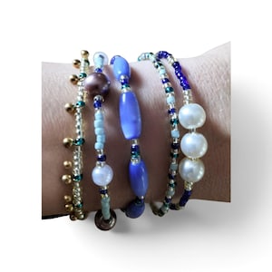 Puede incluir: Un conjunto de cinco pulseras de cuentas con una variedad de formas y colores, incluyendo dijes metálicos dorados, cuentas cilíndricas de color azul claro, azul oscuro y morado, y tres cuentas esféricas blancas grandes en fila.