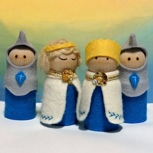 May include: Four felt doll figures, two wearing blue and grey robes and two wearing white robes with gold trim. The figures are standing on a white surface.