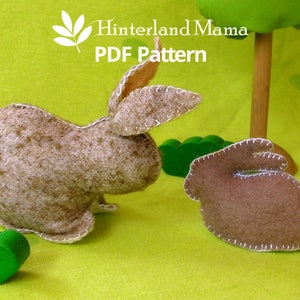 May include: Two brown felt bunny rabbit toys on a green background. The larger bunny is standing on its hind legs with its ears up. The smaller bunny is lying on its side with its ears down. Both bunnies have white felt tails.
