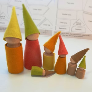 May include: A set of felt gnome dolls in various sizes and colors. The dolls are made of felt and have wooden dowels for bodies. The dolls are wearing felt hats and have felt bodies. The dolls are arranged in a row on a white surface.