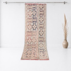 May include: A long, rectangular runner rug hanging from a metal rod. The rug features a repeating geometric pattern in muted tones of blue, brown, beige, and red, with vertical panels separated by thin borders and a fringed edge at the bottom.