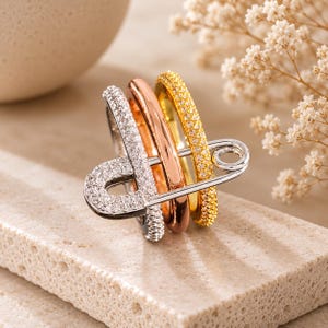 May include: A multi-tone ring featuring three distinct bands in silver, rose gold, and yellow gold tones. The silver band is encrusted with clear stones, the rose gold band is smooth, and the yellow gold band is textured with small clear stones.