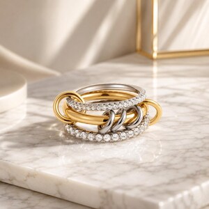 Può includere: Un anello multistrato caratterizzato da due fasce incastonate con pietre chiare, una color argento e una color oro, collegate da una fascia centrale dorata e due piccoli anelli argentati intrecciati che creano una struttura complessa.