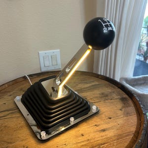 May include: A gear shifter lamp featuring a black spherical knob with a white gear pattern, a metallic silver shaft with integrated warm yellow LED lighting, and a black accordion-style rubber boot mounted on a square chrome base with four screws.