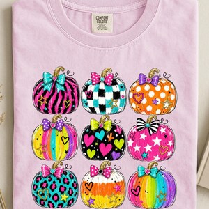 May include: A light pink t-shirt featuring a graphic print of nine pumpkins arranged in a grid. Each pumpkin displays a unique pattern including zebra stripes, polka dots, leopard print, and rainbow colors, accented with glittery bows, hearts, and stars.