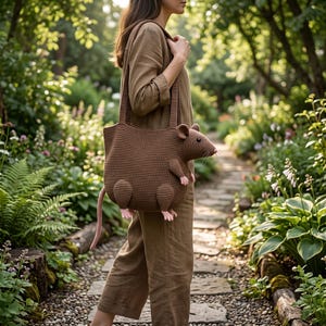 May include: A brown crocheted shoulder bag shaped like a rat, featuring a pointed snout, small round ears, a long tail, and four small paws with pink accents. The bag has a wide shoulder strap and a textured, woven surface throughout the body.