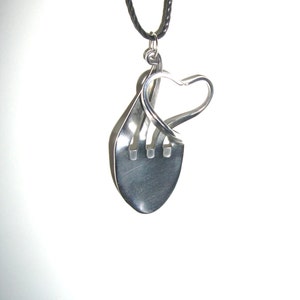 以下が含まれることがあります： フォークをモチーフにした、ハート型のシルバーペンダント。フォークの歯が曲げられてハートを形作り、黒いコードネックレスに吊り下げられています。ペンダントは金属製で、光沢のある仕上がりです。