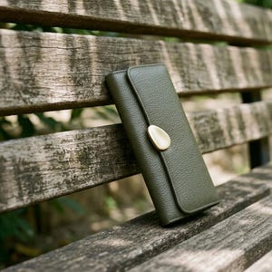 May include: A long, rectangular olive green wallet with a textured, pebbled surface. The wallet features a flap closure secured by a smooth, rounded, gold-toned metal accent piece centred on the front flap, providing a distinct contrast.