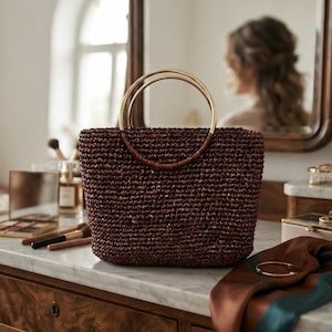 May include: A dark brown woven handbag featuring a structured rectangular body and two circular gold-toned metal handles. The bag has a textured, braided appearance and sits upright, showcasing a consistent deep brown colour throughout its exterior.