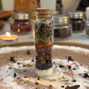 May include: A clear glass vial with a cork stopper, filled with layered ingredients including white salt, dark brown stones, orange-red fragments, green herbs, and a small rolled paper scroll, standing upright in a circle of scattered salt and spices.