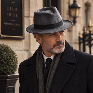 Può includere: Un cappello fedora in feltro grigio con un nastro nero. Il cappello presenta una corona pizzicata e una tesa strutturata. Il materiale ha un aspetto testurizzato e mélange, e il nastro nero circonda la base della corona, creando contrasto.