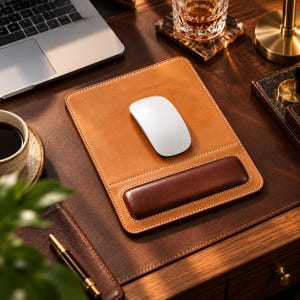 Peut inclure: Un tapis de souris rectangulaire en cuir beige avec une surface lisse et un repose-poignet rembourré marron foncé. Une souris d'ordinateur sans fil blanche est placée au centre du tapis. Le tapis présente des coutures visibles sur les bords.