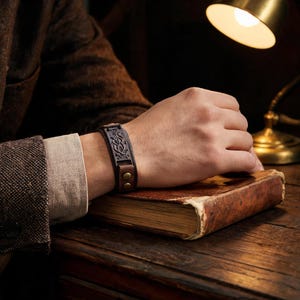 Pode incluir: Uma pulseira de couro castanho escuro com um padrão intrincado de nós celtas em relevo e dois fechos metálicos cor de latão. A pulseira é usada no pulso, apresentando uma superfície texturizada e um design de fecho seguro e ajustável.