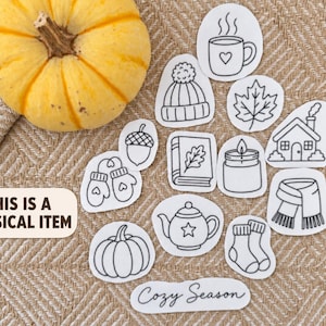 May include: A yellow pumpkin and a collection of white stickers with black line art illustrations of a mug, beanie, leaf, house, acorn, mittens, book, candle, teapot, pumpkin, scarf, and socks, with text reading THIS IS A PHYSICAL ITEM and Cozy Season.